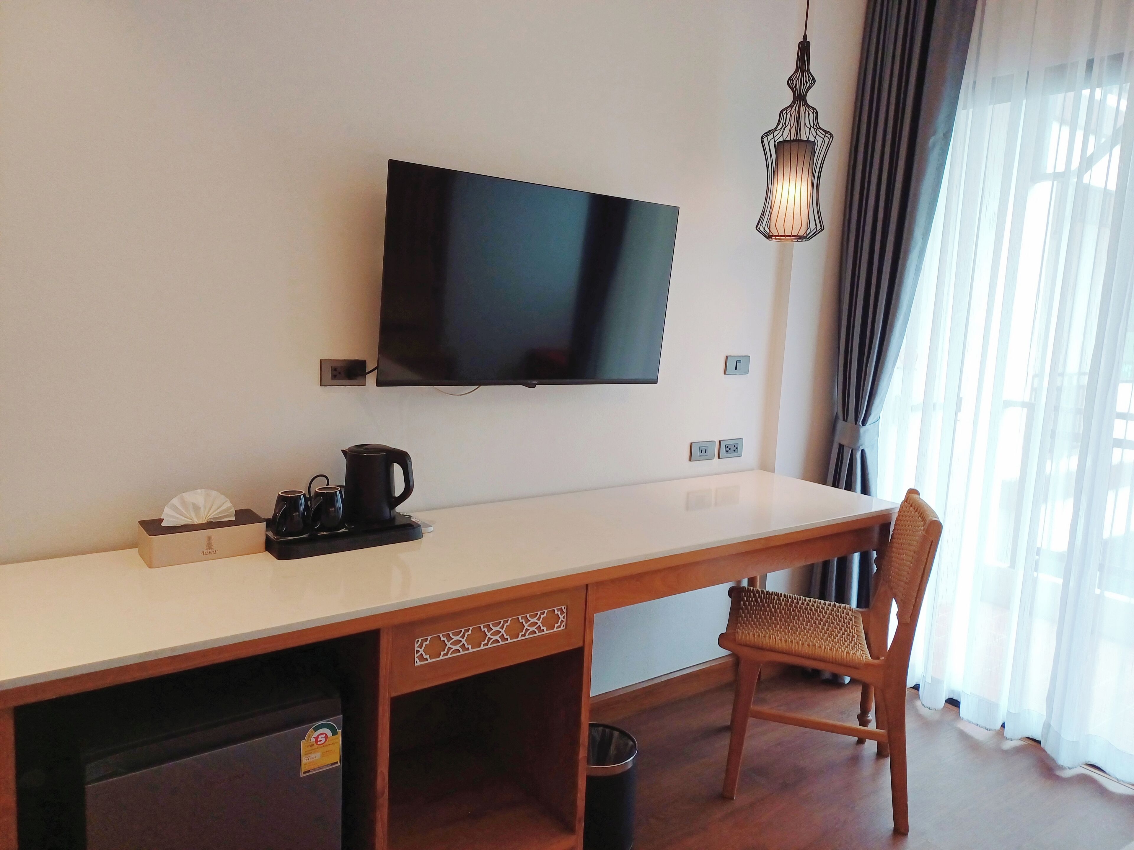 A hotel room with a flat-screen TV mounted on the wall, a wooden desk with a chair, a coffee maker, and a trash bin.