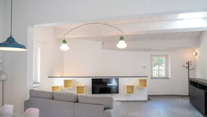 Un salon moderne avec un canapé gris, une table à manger en verre, un téléviseur à écran plat fixé au mur et un plafond avec des poutres apparentes.