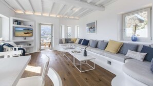 Un salon moderne avec un canapé blanc, une table à manger et un téléviseur fixé au mur. De grandes fenêtres offrent une vue sur l’extérieur.