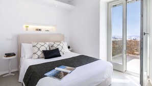 Une chambre moderne avec un grand lit, une table de chevet avec des livres, une commode, une petite étagère avec des objets décoratifs et une vue à travers une porte-fenêtre coulissante.