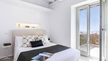 A modern bedroom with a large bed, a bedside table with books, a nightstand, a small shelf with decorative items, and a view through a sliding glass door.