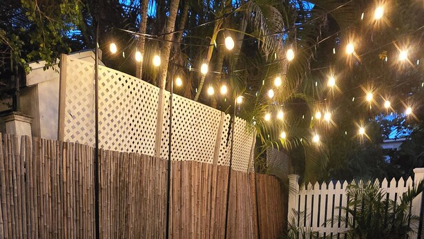 An outdoor dining area with a wooden table and chairs, string lights, and a white picket fence in the background.