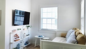 A modern living room with a white sofa, a television mounted on the wall, a dining table with plates and glasses, and a large window with blinds.