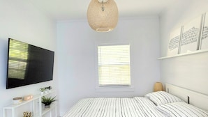 A bedroom with a bed, a TV, a shelf with plants, a window, and a hanging light fixture.