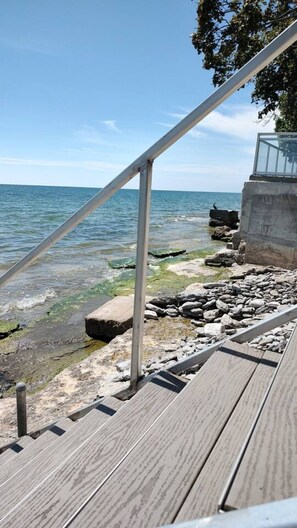 A wooden deck with a metal railing leading to a rocky shoreline by the sea. - Sakura Cottage - Serenity on PEC Waterfront (Prince Edward)