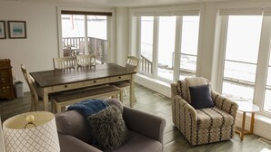 En rummelig opholdsstue med spisebord, to sofaer og store vinduer med udsigt til naturen.