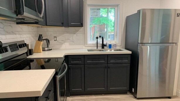 A modern kitchen with dark cabinets, a stainless steel refrigerator, a microwave, and a window with a view of trees. - Two-Bedroom Wellington Apartment (Prince Edward)