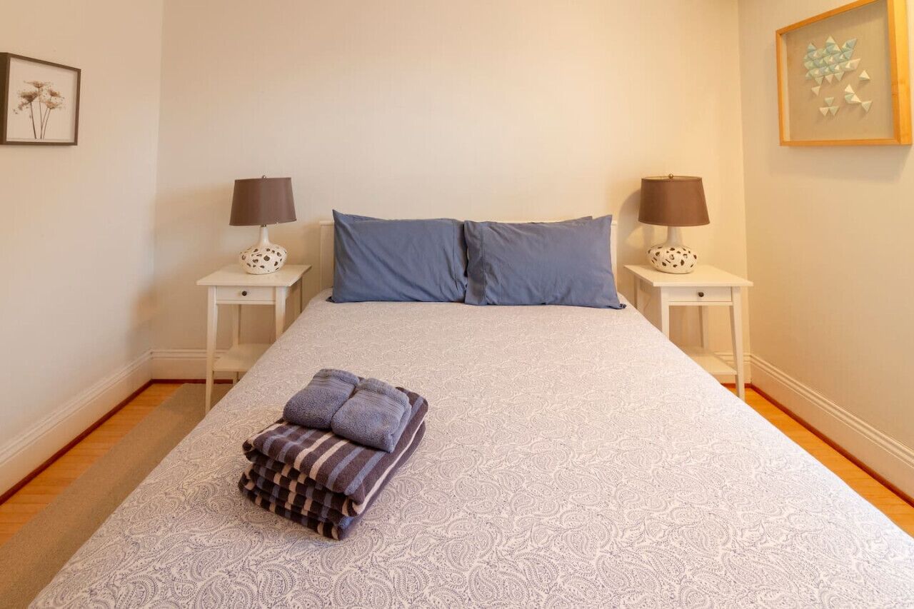Een net opgemaakt bed met een geboeide spreidekbedovertrek, twee kussens en een opgevouwen handdoek op een witte nachtkastje.