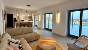 A modern living room with a sofa, coffee table, and chair, a kitchen in the background, and large windows with a view of the sea. - Faragas Hill (Paros Island)