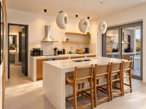 A modern kitchen with a central island, wooden chairs, pendant lights, and a view through a glass door. - Faragas Hill (Paros Island)