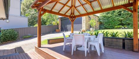 An outdoor patio with a wooden deck, white plastic chairs, and a covered seating area with a corrugated metal roof.