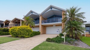 A modern two-story residential building with a covered driveway, a balcony, and a well-maintained garden with palm trees and shrubs.
