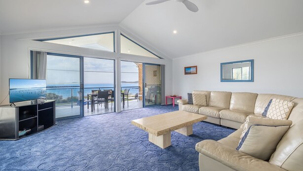 A spacious living room with a large sofa, a coffee table, and a TV on a stand. There is a balcony with a view of the sea and a dining area with chairs and a table.