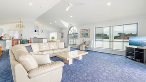 A spacious living room with a large sectional sofa, a coffee table, and a television. The room has a high ceiling with a fan, a large window with a view, and a kitchen area in the background.