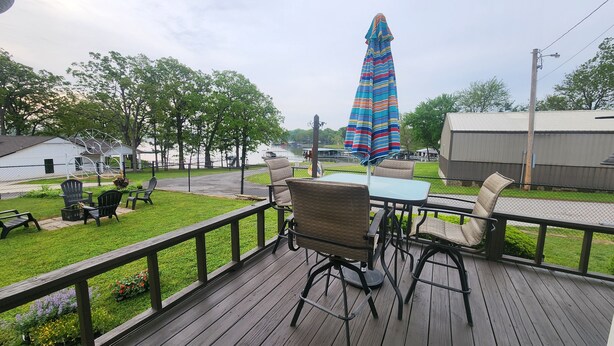 A wooden deck with outdoor furniture, a table, and chairs, overlooking a grassy area with a fence and a view of a body of water and buildings in the distance. - Lakeview, 4 beds, boat parking & ramp, at Red Arrow Marina (Afton)