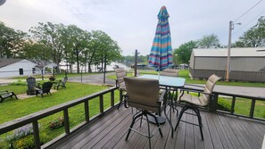 Une terrasse en bois, agrémentée de mobilier d’extérieur, d’une table et de chaises, donnant sur une zone herbeuse délimitée par une clôture, et offrant une vue sur une étendue d’eau et des bâtiments situés au loin.