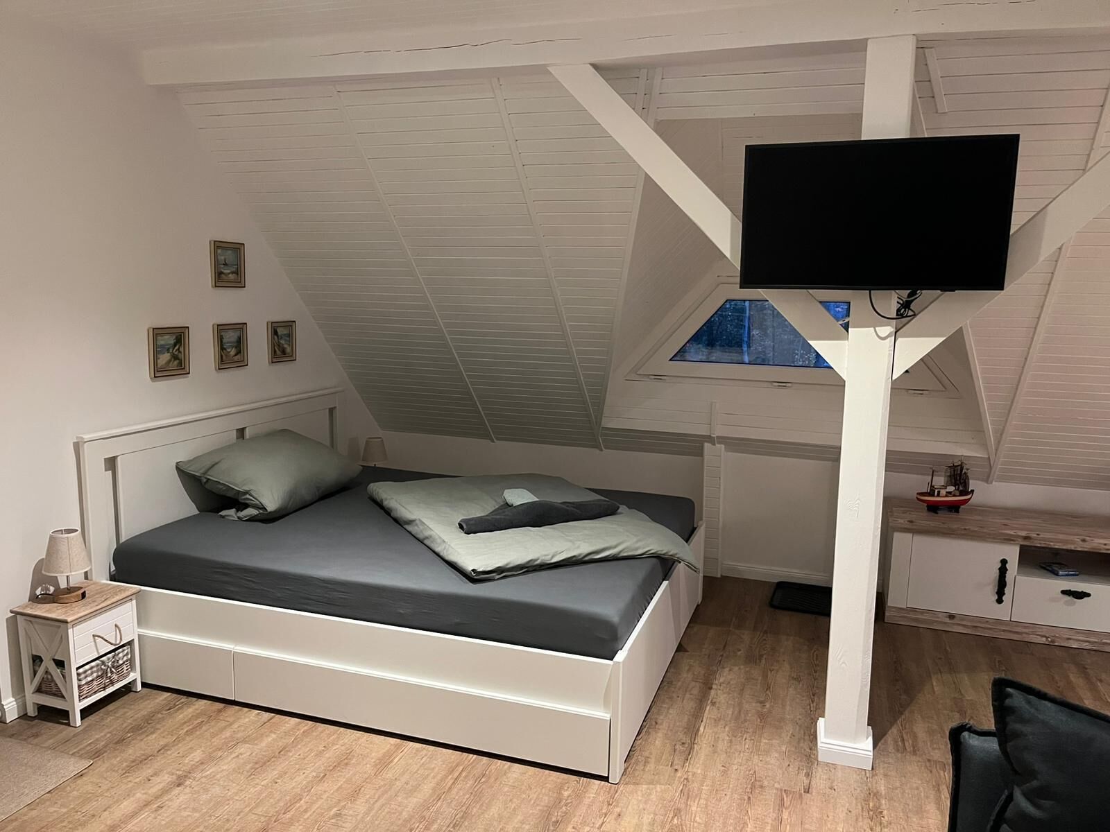 Ein Schlafzimmer mit schrägen Wänden, einem Bett mit grauer Bettwäsche, einem weißen Nachttisch, einer kleinen Kommode und einem an der Wand befestigten Fernseher.