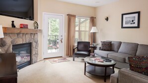 A living room with a stone fireplace, a television mounted above it, a sofa, a chair, a coffee table, and a door leading to an outdoor area with a balcony. - Chalet 98 at the Official Collins Lake Resort (Government Camp)