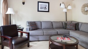 A living room with a grey sofa, a brown leather chair, a glass coffee table with remote controls, and a framed picture on the wall. - Chalet 98 at the Official Collins Lake Resort (Government Camp)