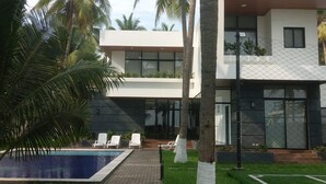 Een modern huis met een zwembad, witte ligbedden en palmbomen.