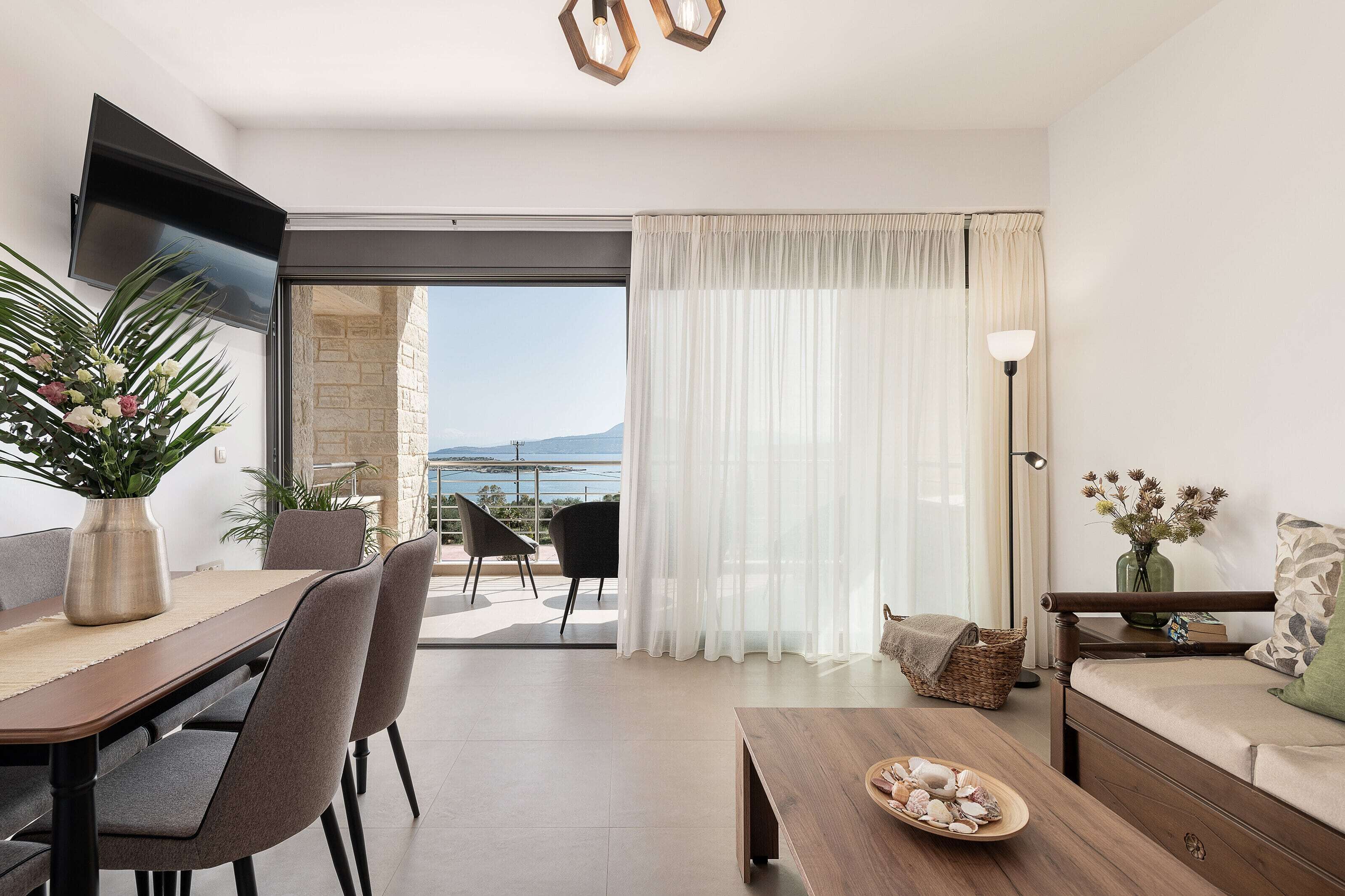 Un salon moderne comprenant un coin repas, un canapé, un téléviseur fixé au mur et une vue sur la mer grâce à de grandes fenêtres.