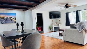 A spacious living room with a wooden ceiling, a dining area with a table and chairs, a fireplace, a television, and a large window with curtains.