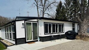 A mobile home with a number 130 sign, large windows, a covered grill, and a gravel driveway.