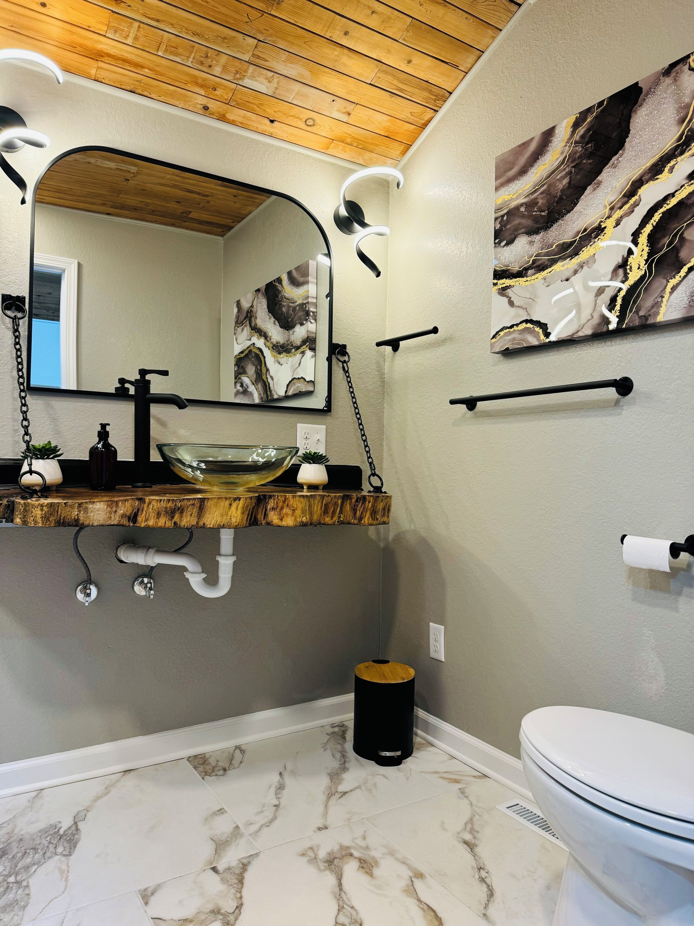 Une salle de bain avec un plafond en bois, un sol carrelé de marbre, un lavabo en verre, un robinet noir, des toilettes et une œuvre d’art murale en marbre.