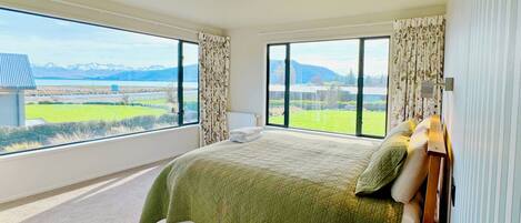 A bedroom with a large bed, a wooden nightstand, and a view of a green landscape through large windows.