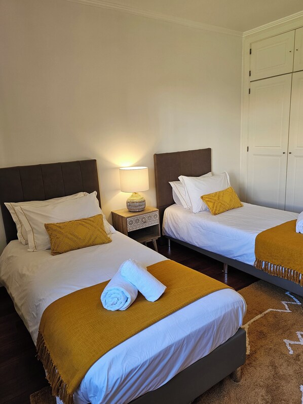 A hotel room with two beds, each with white bedding and a brown headboard. There is a nightstand with a lamp between the beds. - Villa Jarros Private Pool & Golf (Aroeira)
