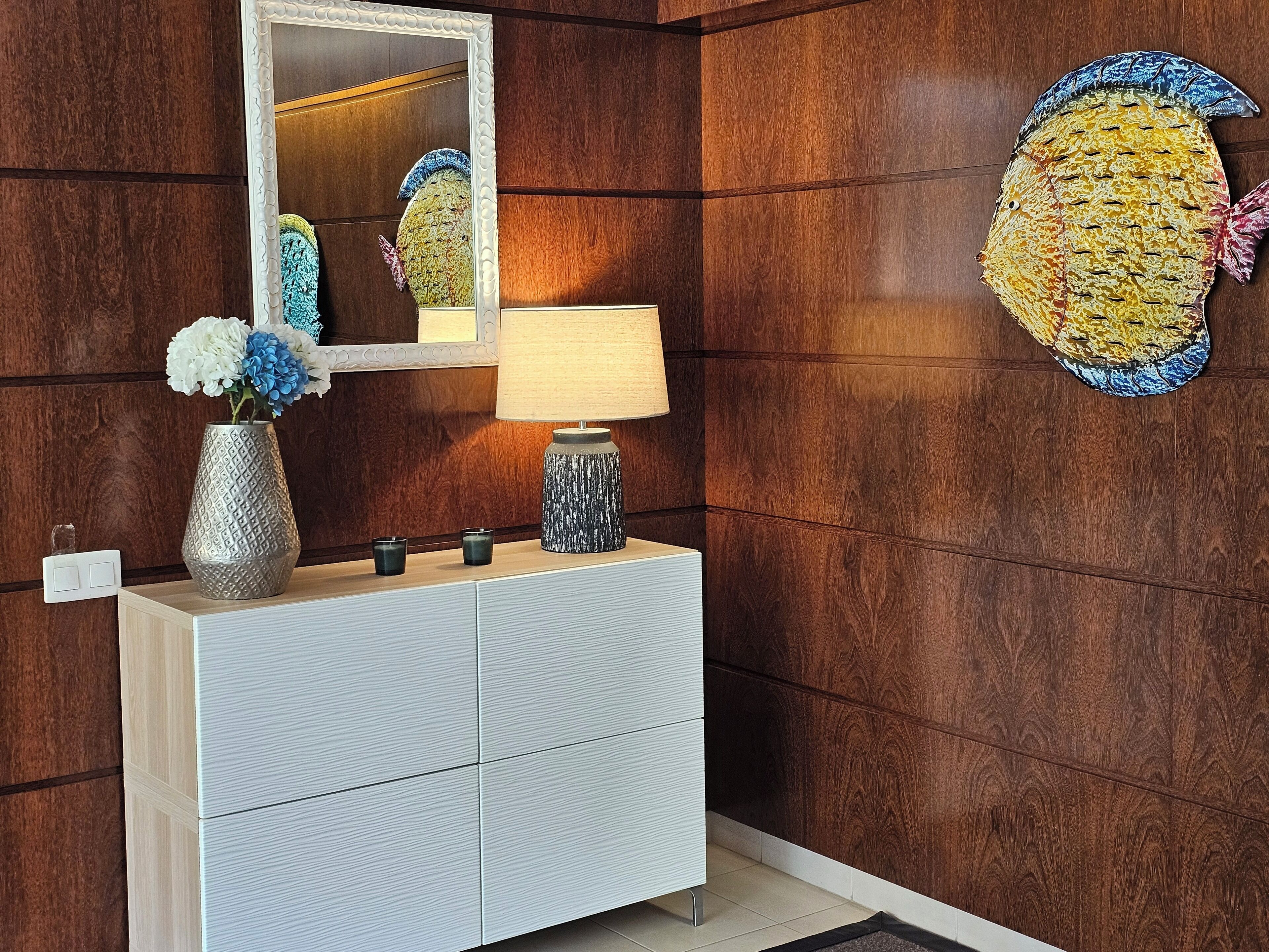 Een moderne badkamer met een houten wastafel, een spiegel, een vaas met bloemen, een lamp en een decoratief wandobject.