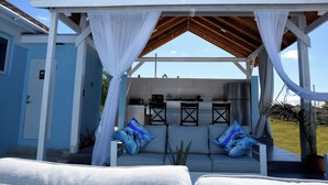 A covered outdoor seating area with a kitchen in the background, white curtains, and blue cushions. - Glory of Eden (Nassau)