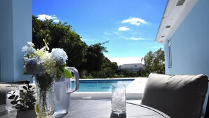 A table with a vase of flowers, a pitcher, and a glass of ice water, overlooking a swimming pool and greenery. - Glory of Eden (Nassau)
