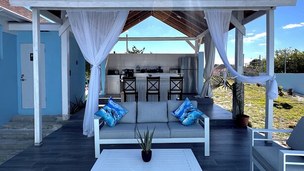 A covered outdoor seating area with a sofa and chairs, a small table, and a kitchen in the background. - Glory of Eden (Nassau)