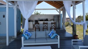 A covered outdoor seating area with a sofa and chairs, a kitchen area with a bar, and a view of the surrounding landscape. - Glory of Eden (Nassau)