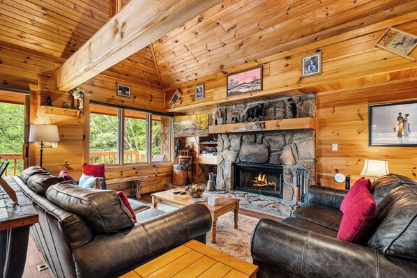 A cozy cabin interior with a stone fireplace, leather sofas, wooden walls, and a large window overlooking greenery. - Dancing in the Moonlight is the Perfect Romantic Getaway (GATLINBURG)