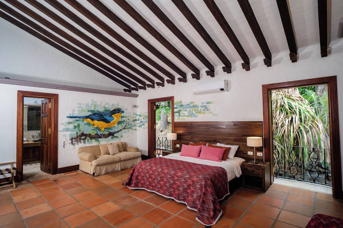 A bedroom with a wooden ceiling, a bed with a red blanket, a beige sofa, and a mural on the wall.