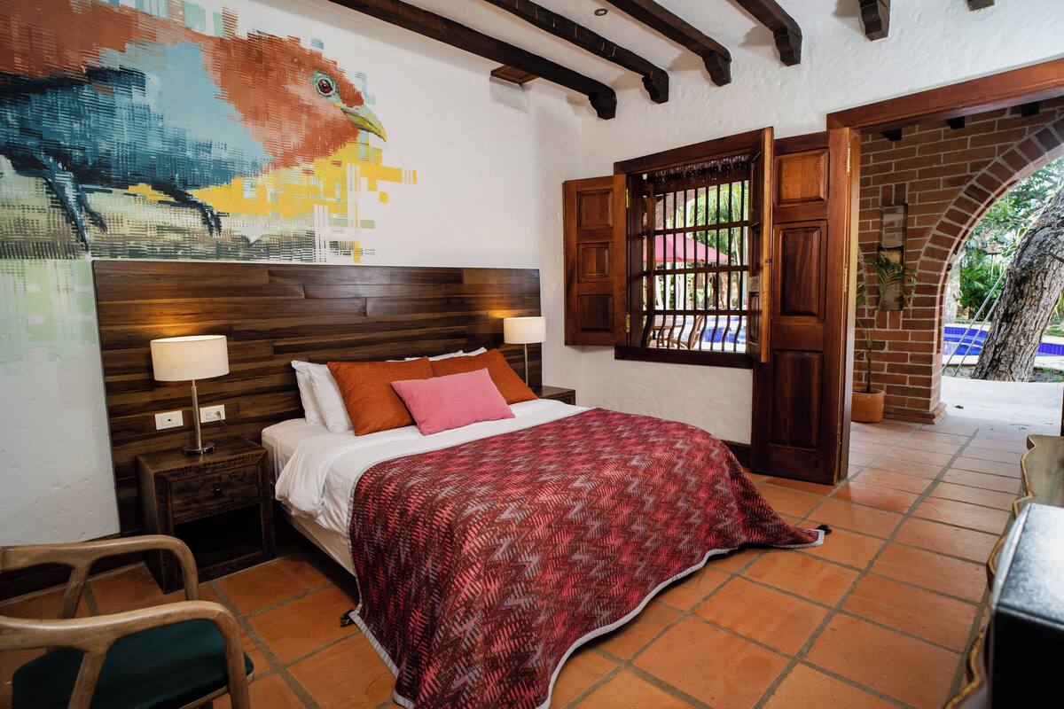 A bedroom with a wooden headboard, a patterned bedspread, two bedside lamps, and a mural on the wall.