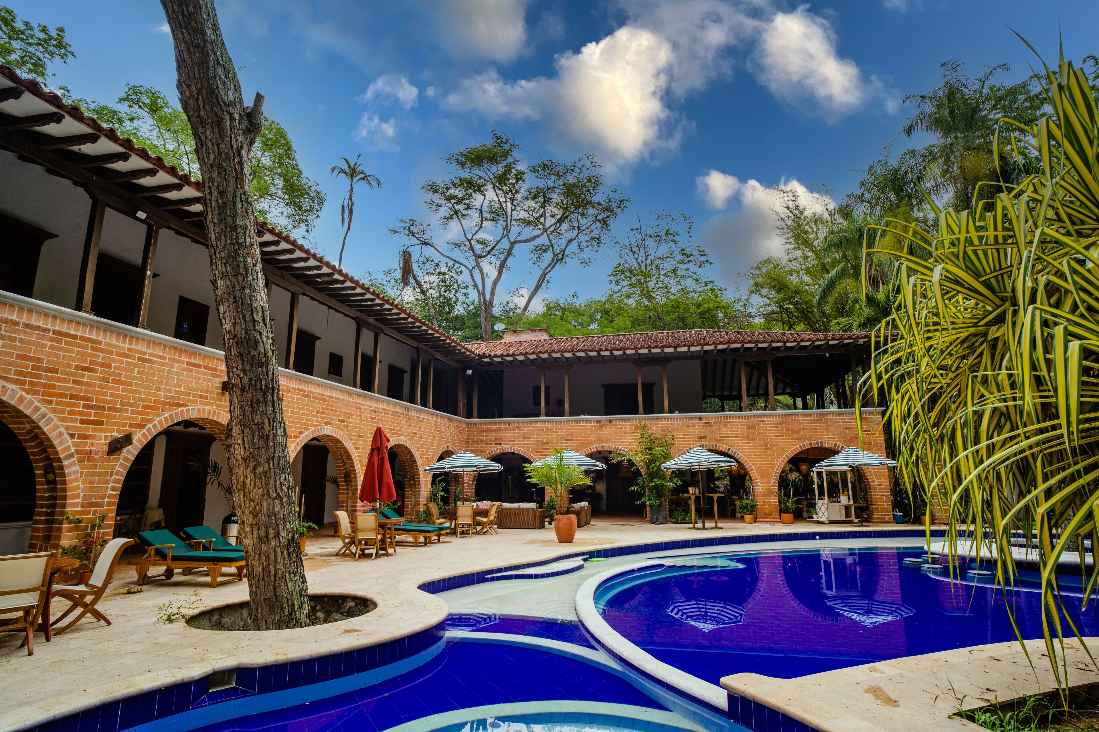 A pool area with a brick building, lounge chairs, and umbrellas.