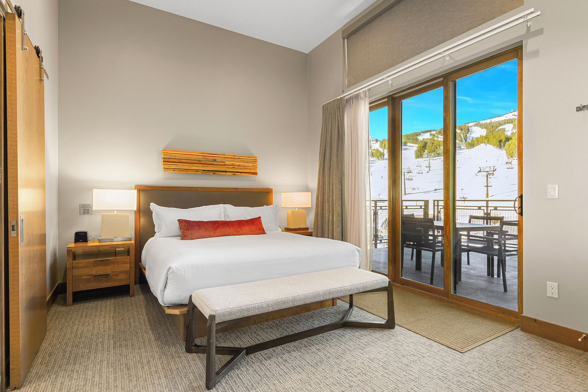 A hotel room with a bed, bedside tables, a bench, a view of a snowy landscape through a sliding glass door.