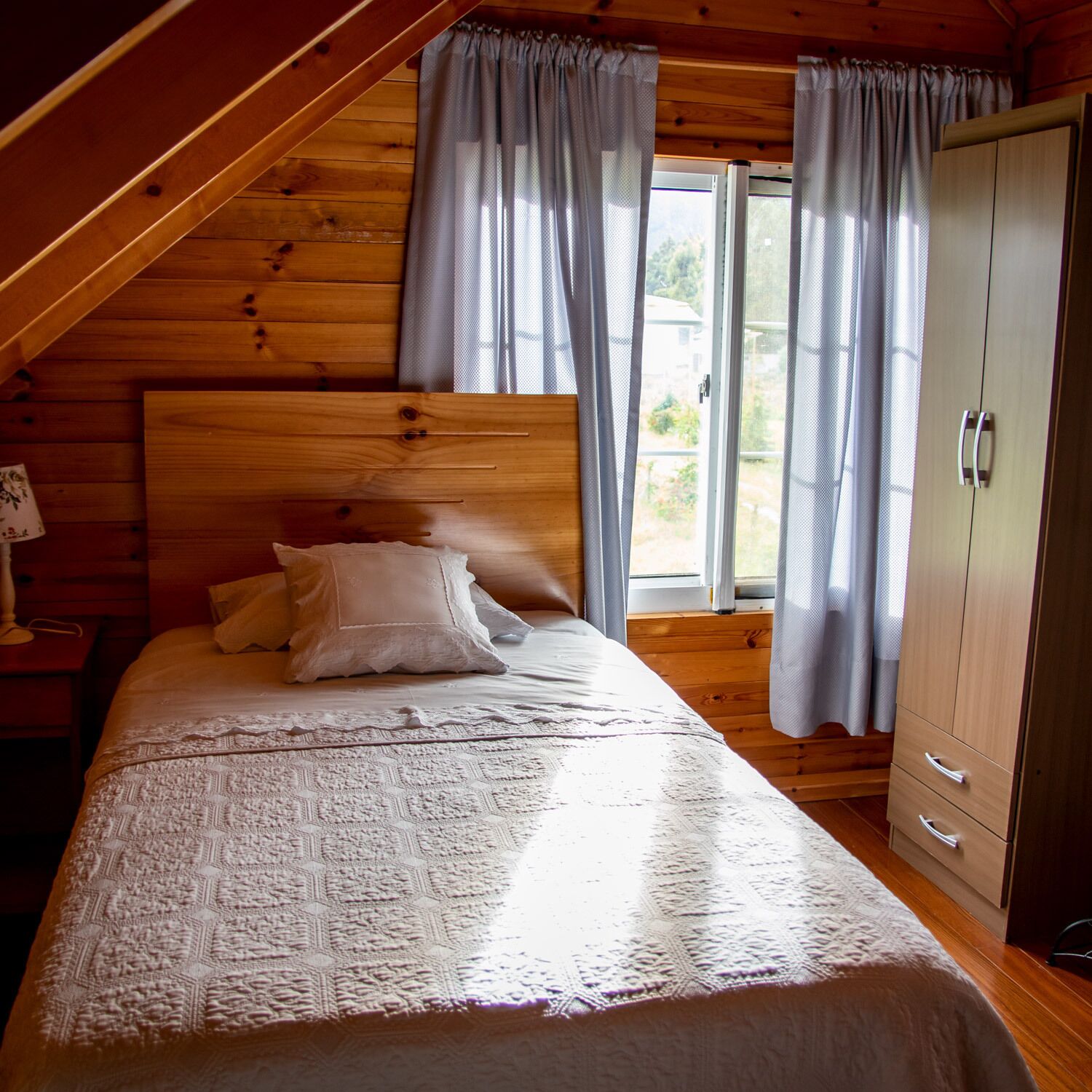 A bedroom with a wooden ceiling, a bed with a patterned bedspread, two pillows, a nightstand with a lamp, and a wardrobe.