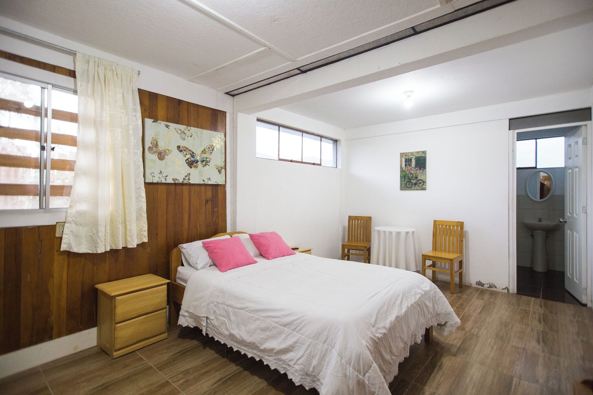 Un dormitorio con una cama, dos sillas, una mesita de noche de madera, una pequeña mesa y una ventana con cortinas.
