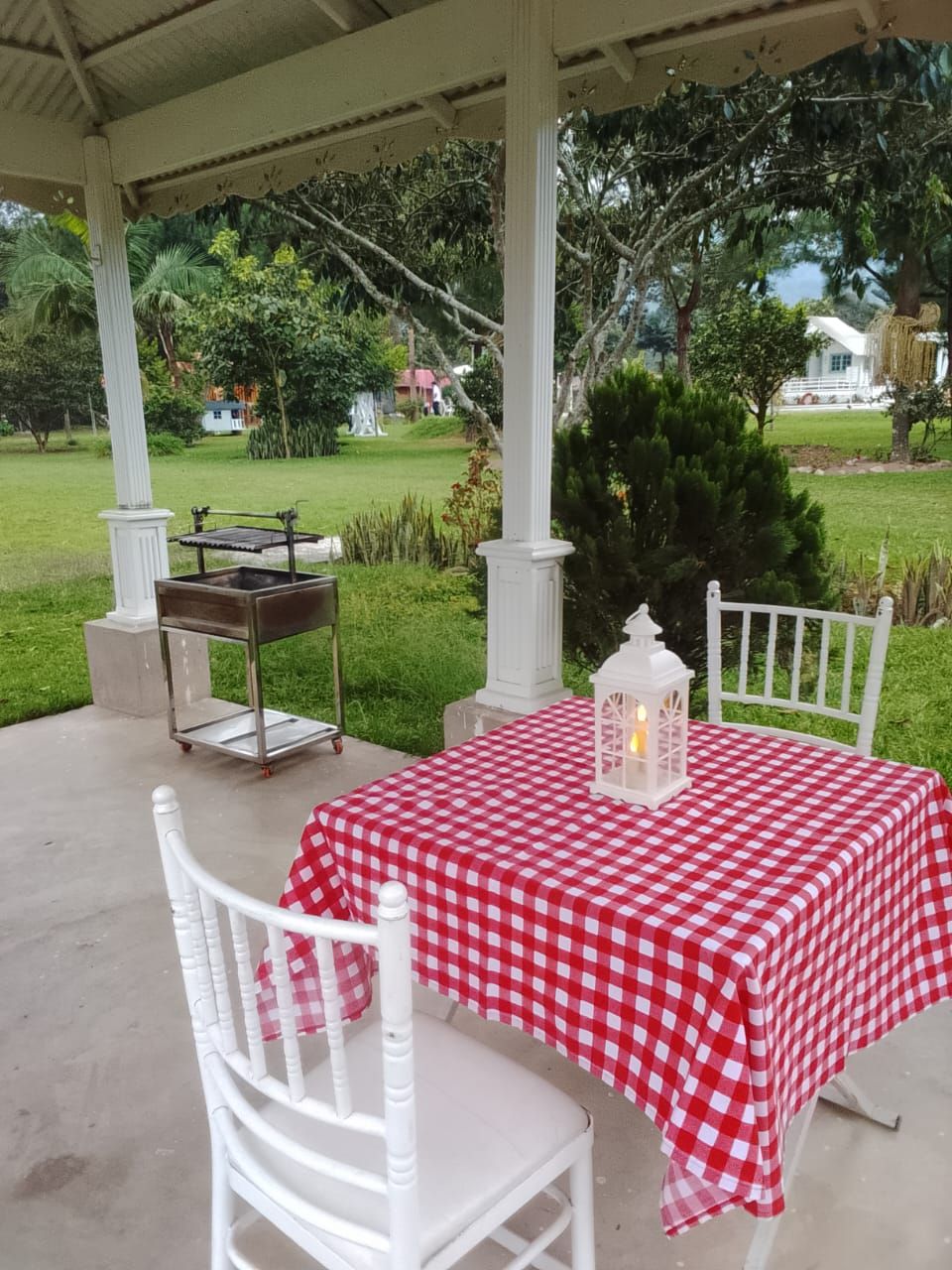 A covered outdoor seating area with a checkered tablecloth, white chairs, and a lantern.