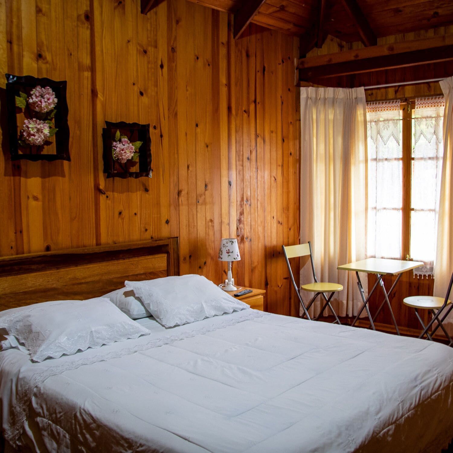 Quarto de madeira com uma cama, uma cadeira, uma mesinha e uma janela com cortinas.