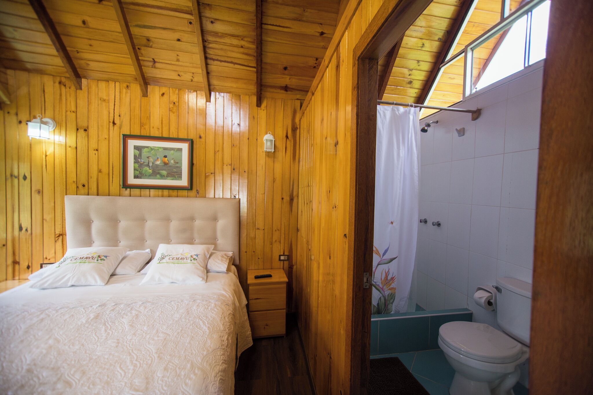 Una habitación con paneles de madera, una cama, una mesita de noche, un cuadro enmarcado y un baño con inodoro y cortina de ducha.