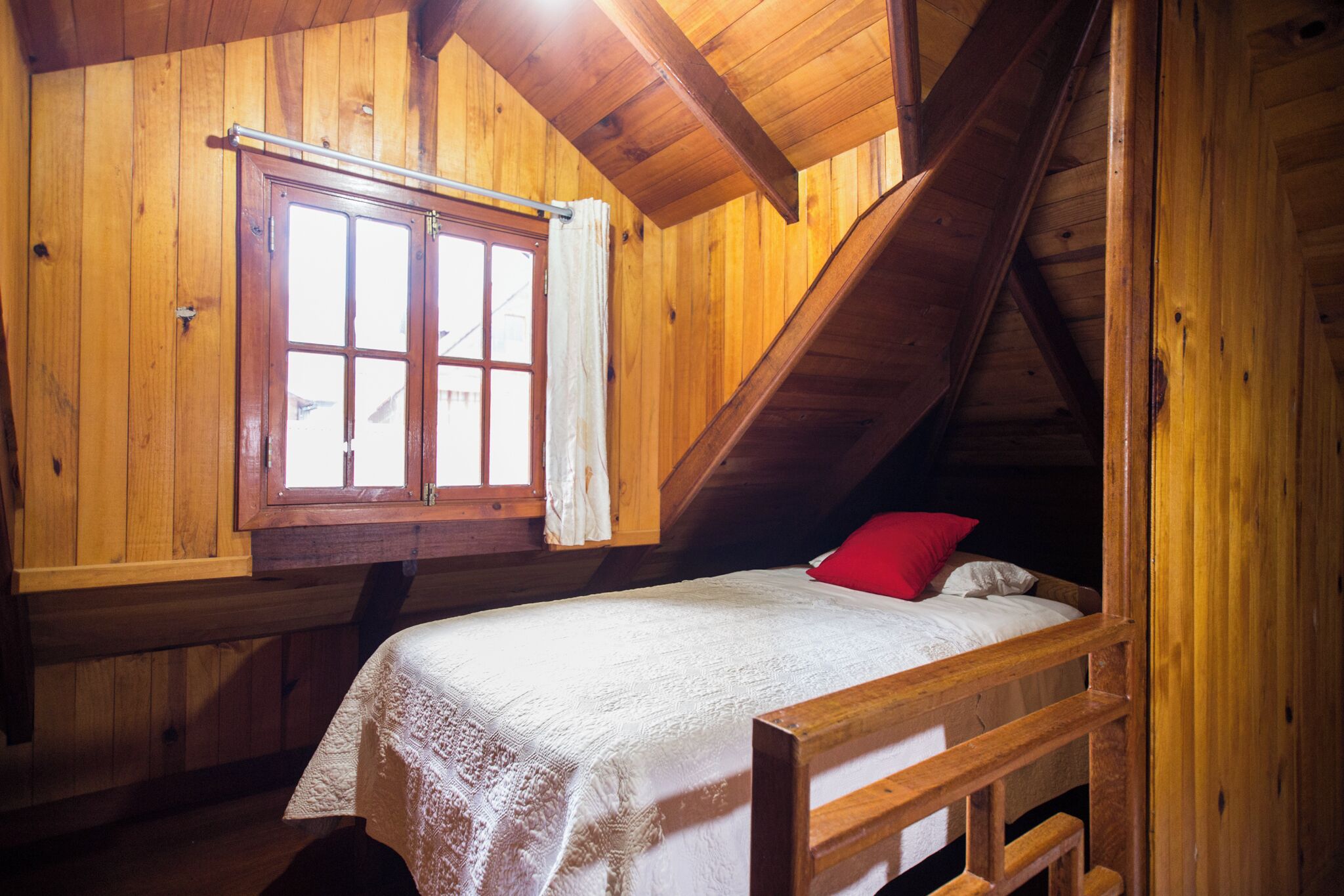 Una habitación con piso de madera, una cama, una ventana con cortinas y techo inclinado.