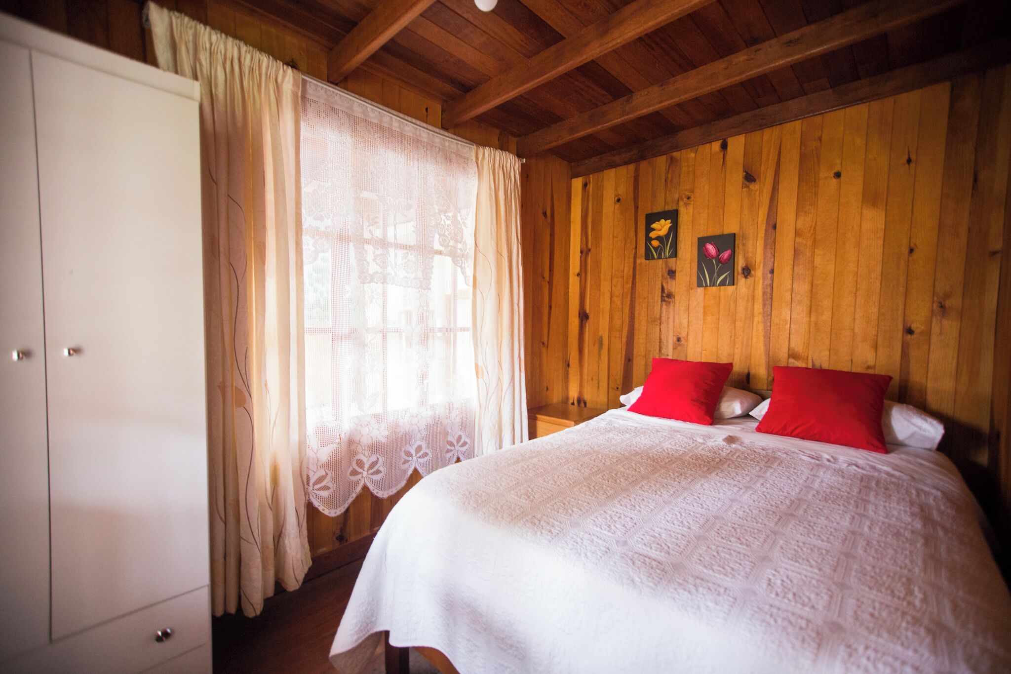 Une pièce aux murs lambrissés, avec un lit, des rideaux blancs et un coussin rouge.