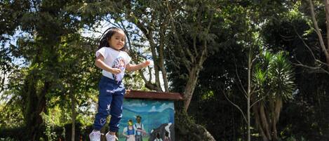 A child jumping on a trampoline in a park with trees and a mural in the background.