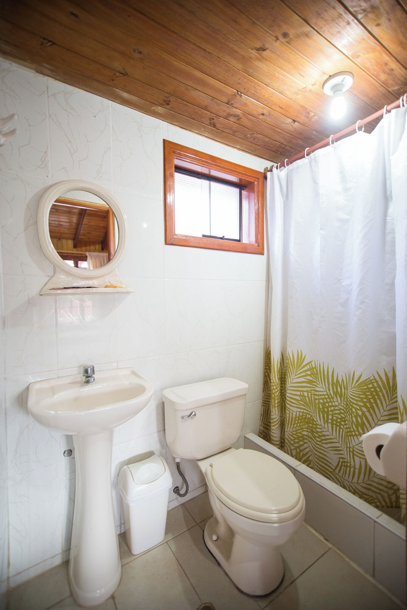 Un baño con techo de madera, lavabo pedestal, inodoro y cortina de ducha con estampado de hojas.