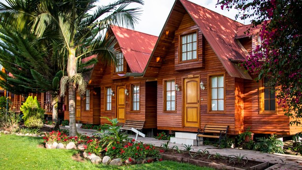 Una hilera de cabañas de madera con techo inclinado, una palmera y un jardín con flores rojas.